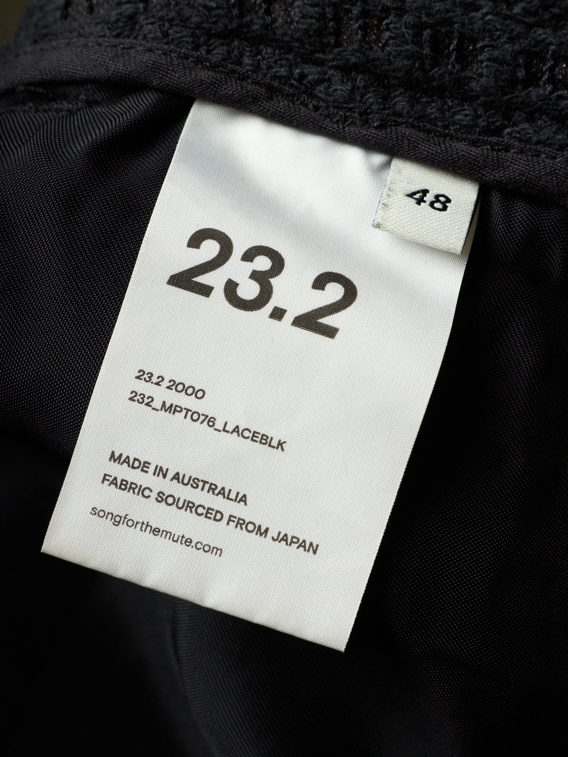Close-up of a black garment interior with a white size tag reading 23.2 and 48, plus text noting made in Australia and fabric sourced from Japan.