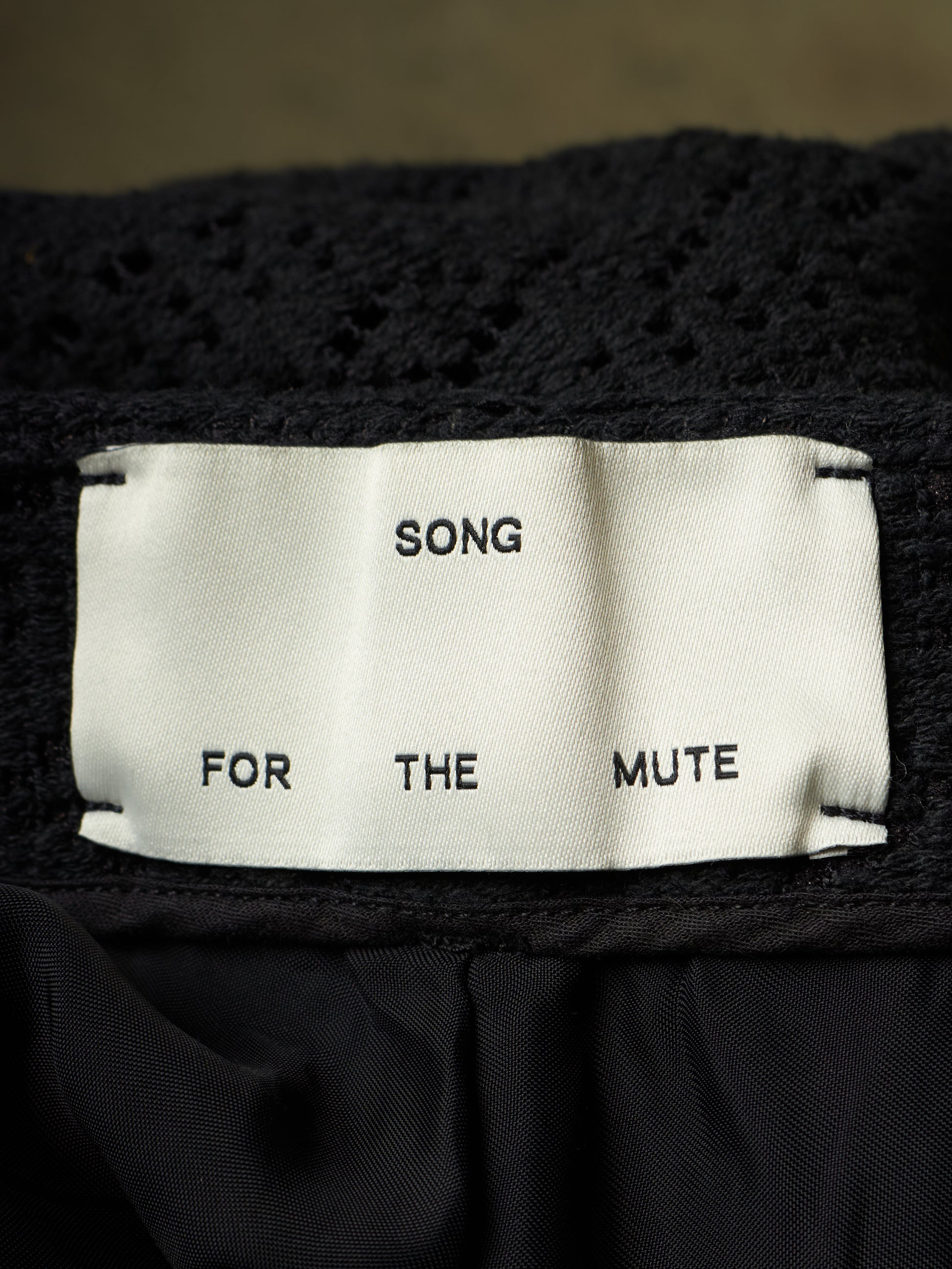 Close-up of a black knit garment with a white fabric label reading 'SONG FOR THE MUTE'.