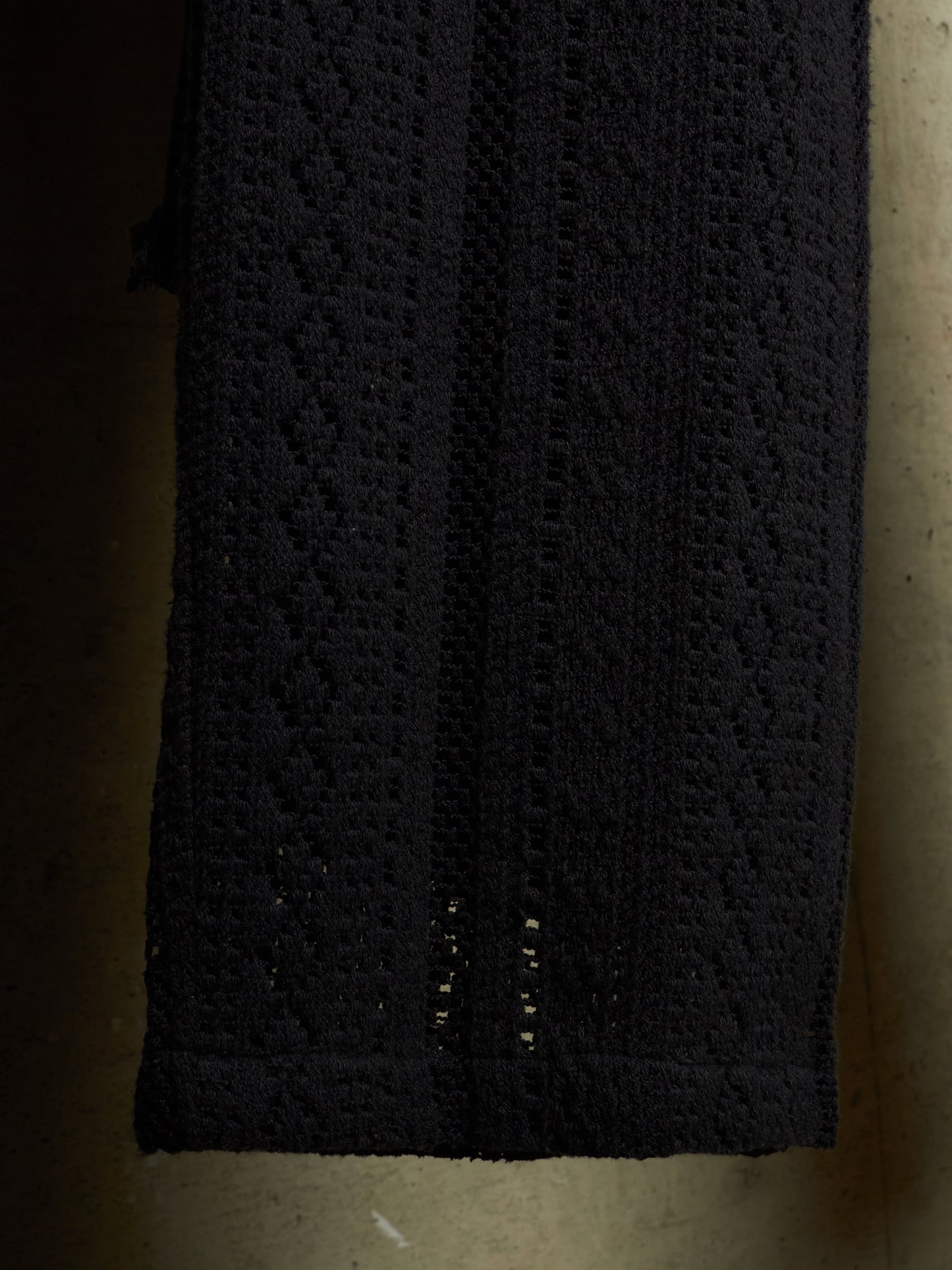 A close-up of a black textured knit fabric, showing a diamond-patterned crochet or lace design with openwork details near the bottom edge.