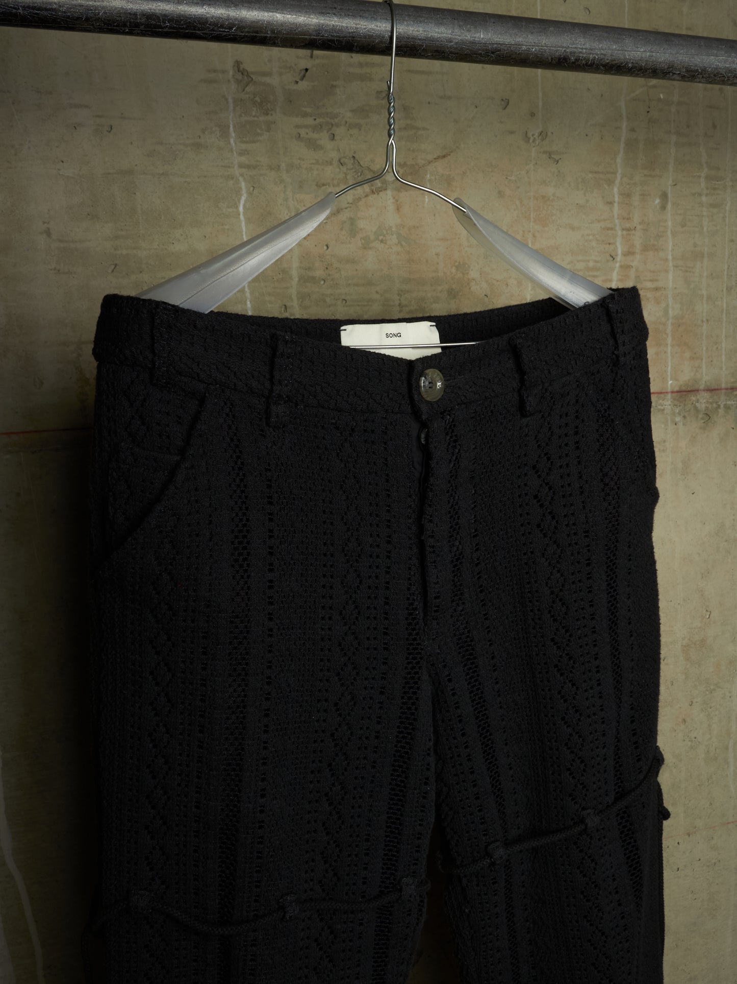 Black cable-knit cropped pants hanging on a metal hanger against a textured concrete wall.
