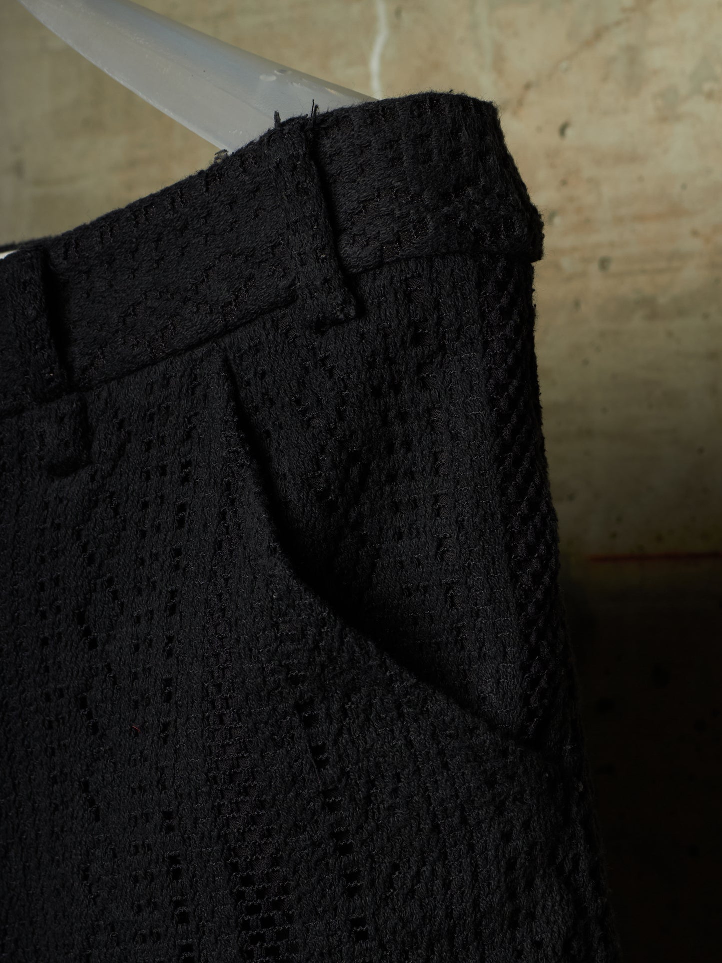 Close-up of a black textured woven fabric garment, showing the waistband and side pocket detail against a warm neutral background.