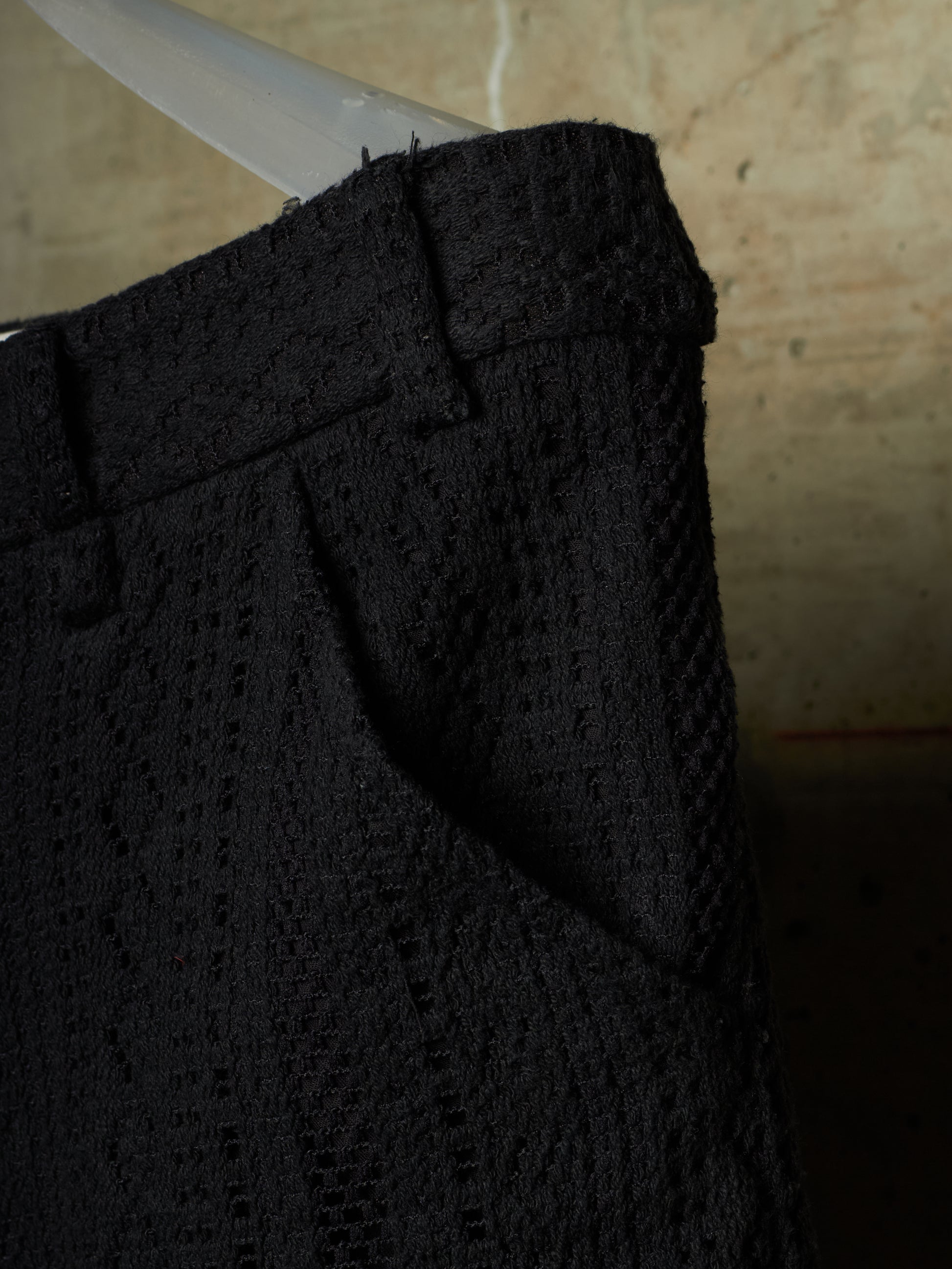 Close-up of a black textured woven fabric garment, showing the waistband and side pocket detail against a warm neutral background.