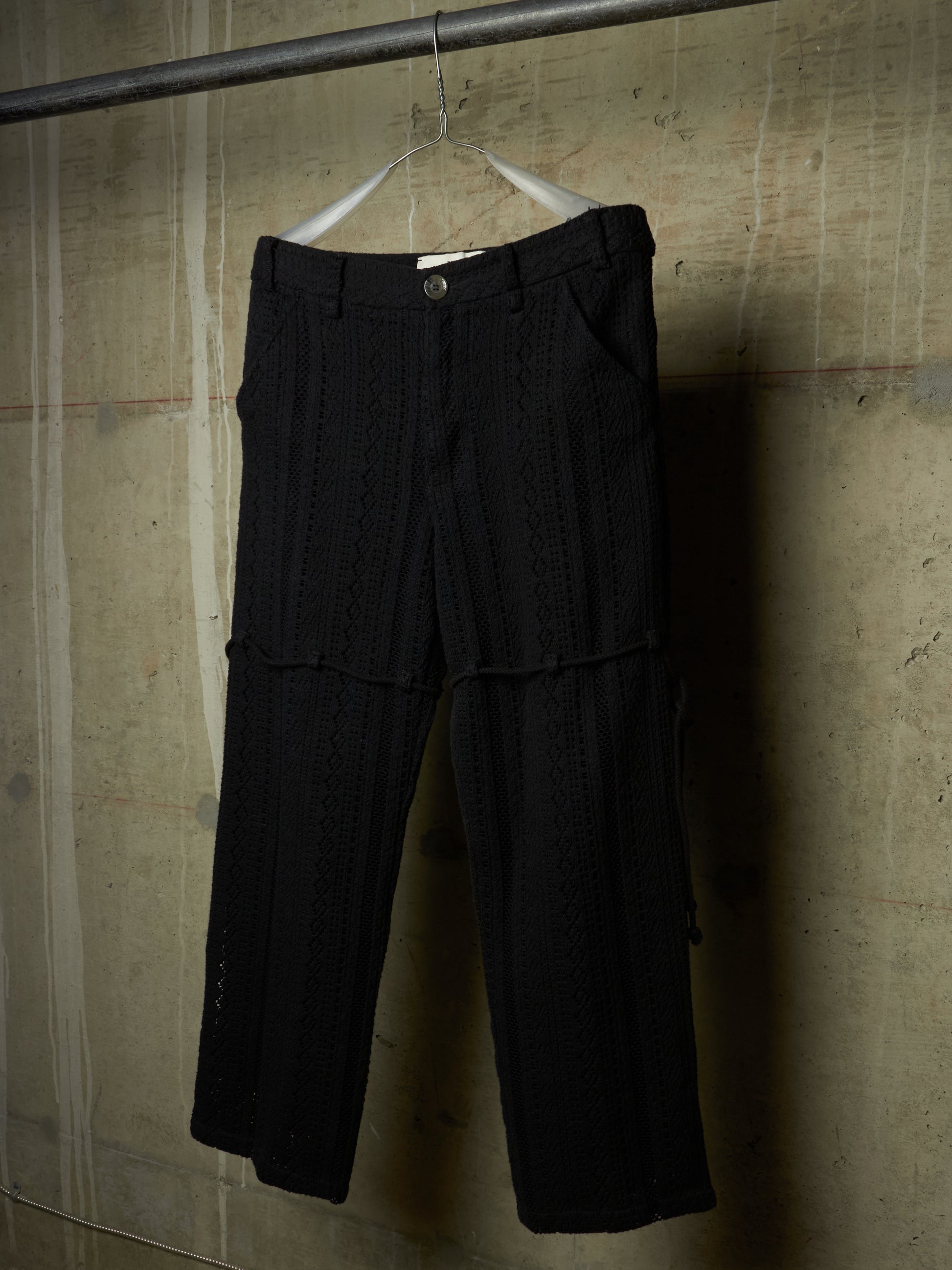 Black textured knit cropped pants hanging on a white hanger from a metal rod against a concrete wall.