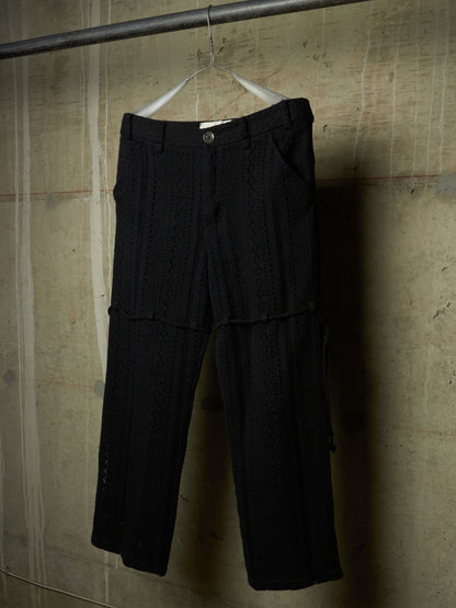 Black textured knit cropped pants hanging on a white hanger from a metal rod against a concrete wall.