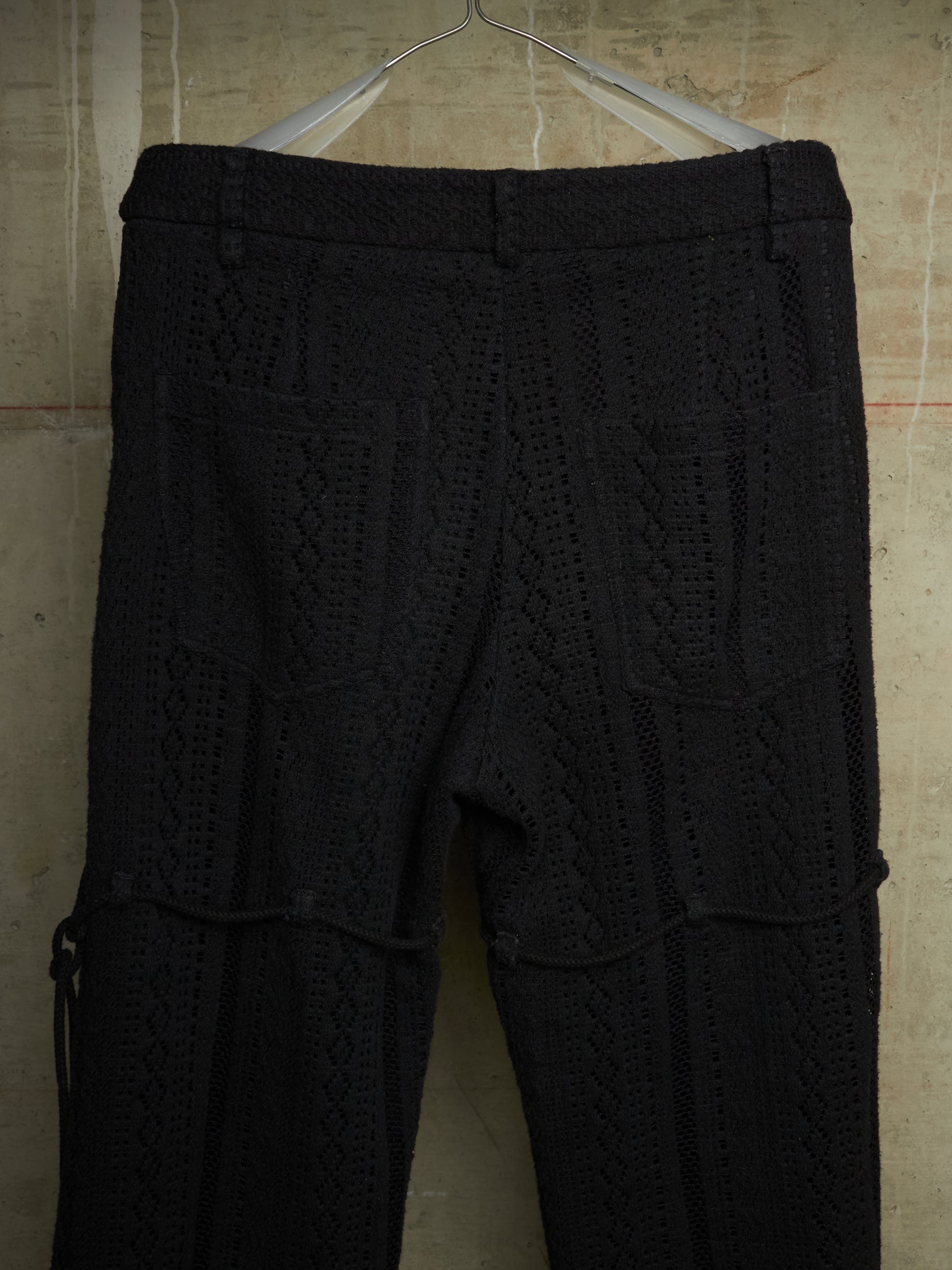 Black textured knit pants with a cable-knit pattern, hanging on a metal hanger against a beige tiled wall.