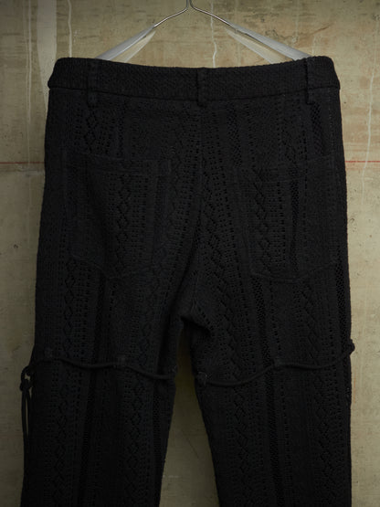 Black textured knit pants with a cable-knit pattern, hanging on a metal hanger against a beige tiled wall.