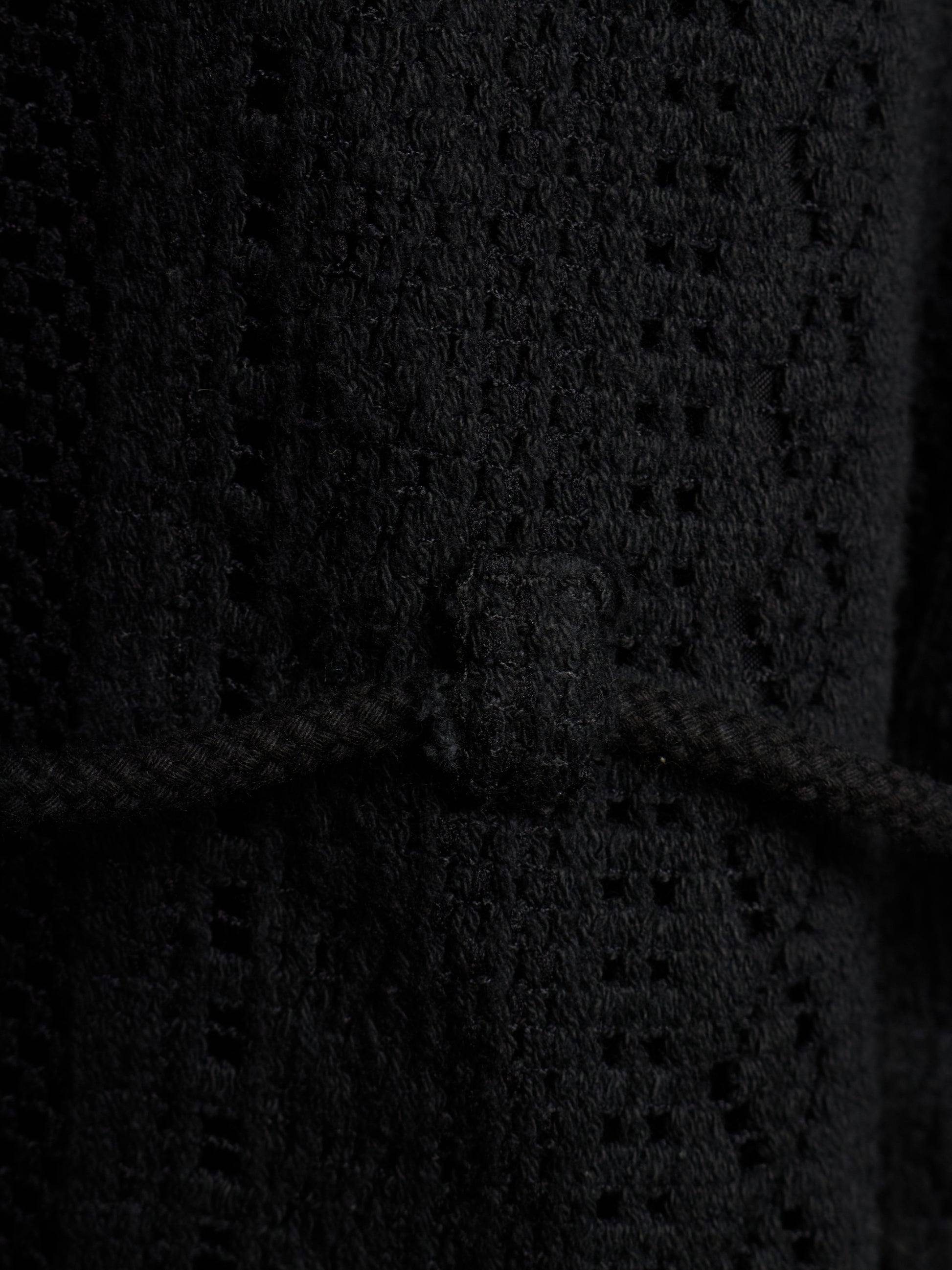 Close-up of a black textured knit garment with a braided cord detail and a small rectangular tab at the center.