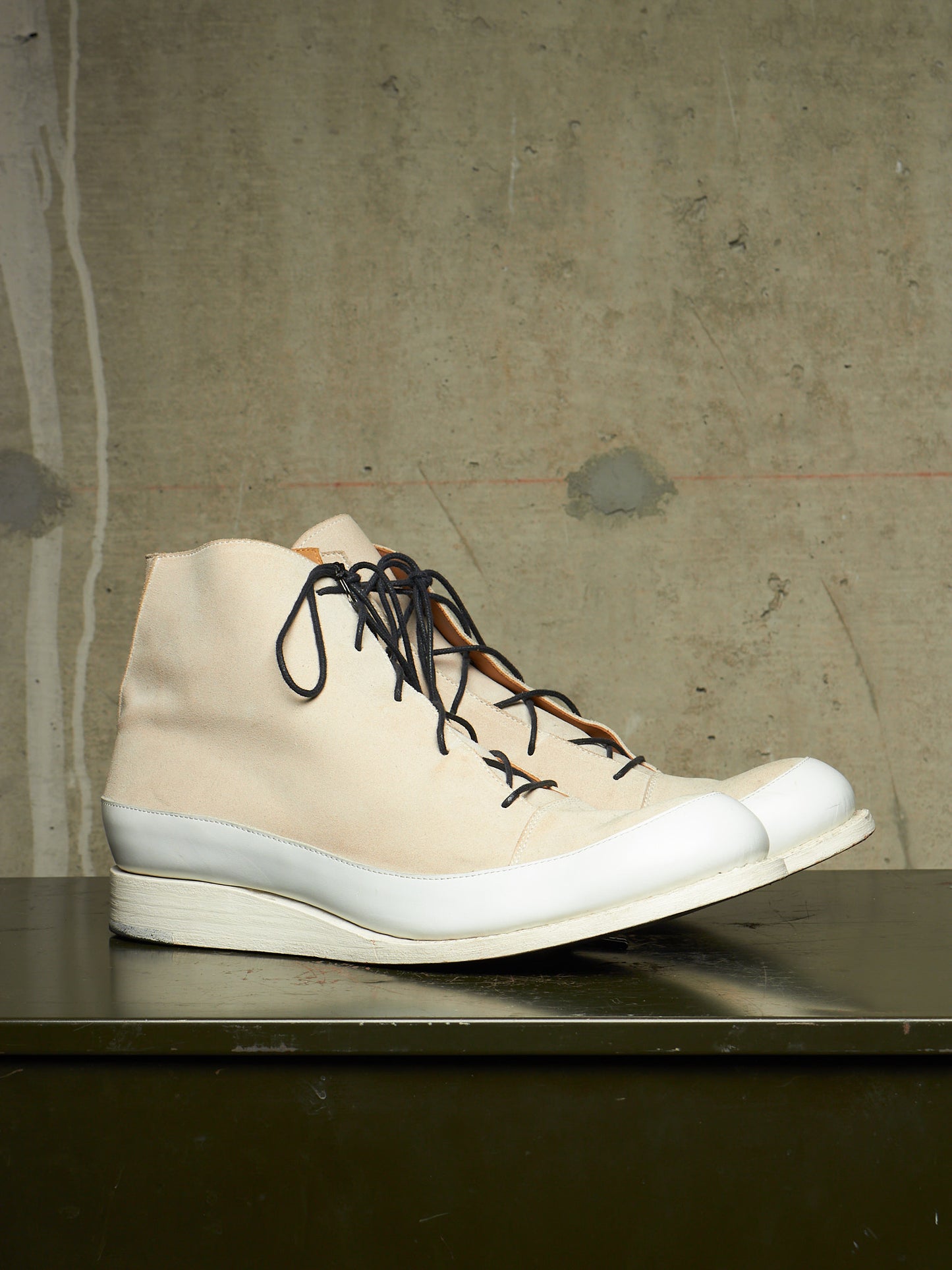 A single high-top sneaker boot in off-white canvas with black laces and a thick white sole, resting on a dark metallic surface against a textured concrete wall.