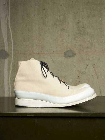 A single high-top sneaker boot in off-white canvas with black laces, a thick white midsole, and a white rubber toe cap, displayed on a dark shelf against a textured concrete wall.