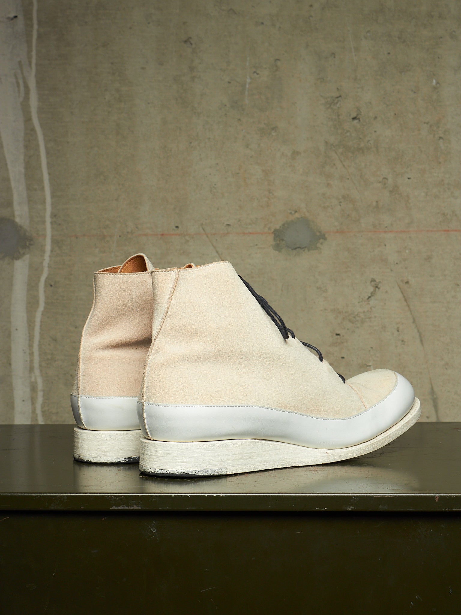 A pair of mid-calf lace-up boots in a light beige and off-white colorway, shown from the side on a dark tabletop against a textured concrete wall.