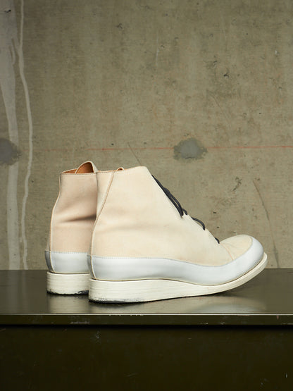 A pair of mid-calf lace-up boots in a light beige and off-white colorway, shown from the side on a dark tabletop against a textured concrete wall.
