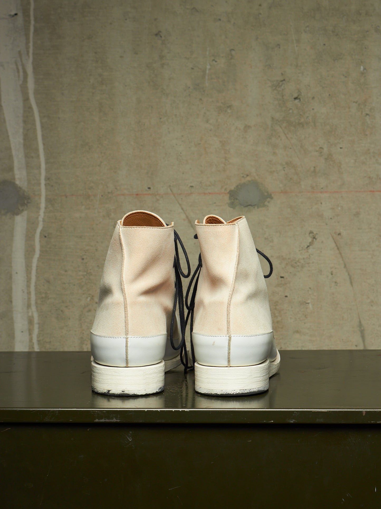 A pair of high-top canvas sneakers viewed from the back, featuring a light beige upper, a white rubber midsole, and black laces, resting on a dark surface against a textured concrete wall.