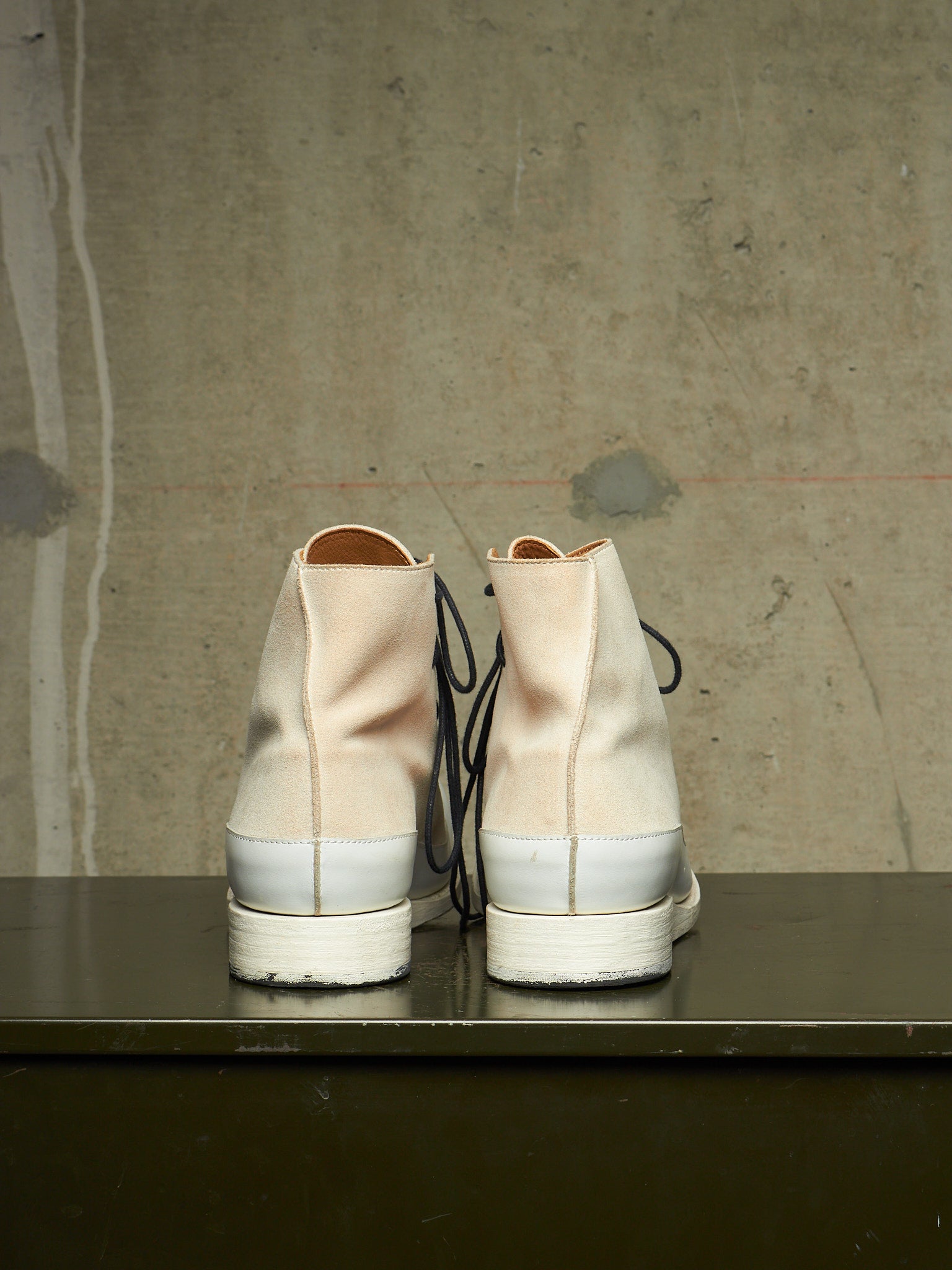 A pair of high-top canvas sneakers viewed from the back, featuring a light beige upper, a white rubber midsole, and black laces, resting on a dark surface against a textured concrete wall.