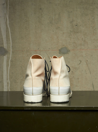 A pair of high-top canvas sneakers viewed from the back, featuring a light beige upper, a white rubber midsole, and black laces, resting on a dark surface against a textured concrete wall.