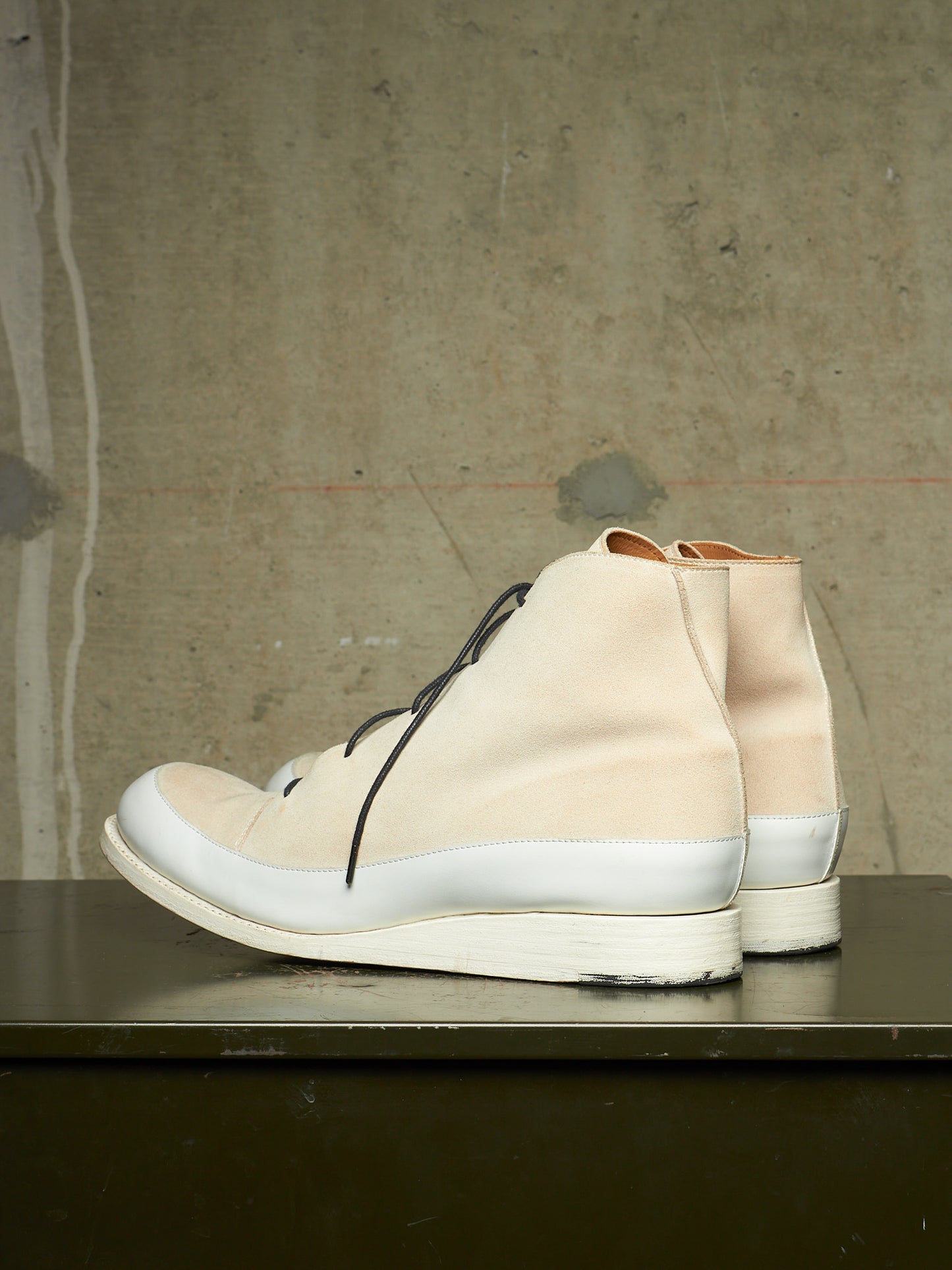 A pair of high-top lace-up sneakers in a light beige and white colorway, displayed on a dark surface against a textured concrete wall.