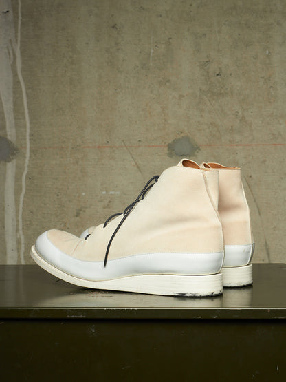 A pair of high-top lace-up sneakers in a light beige and white colorway, displayed on a dark surface against a textured concrete wall.