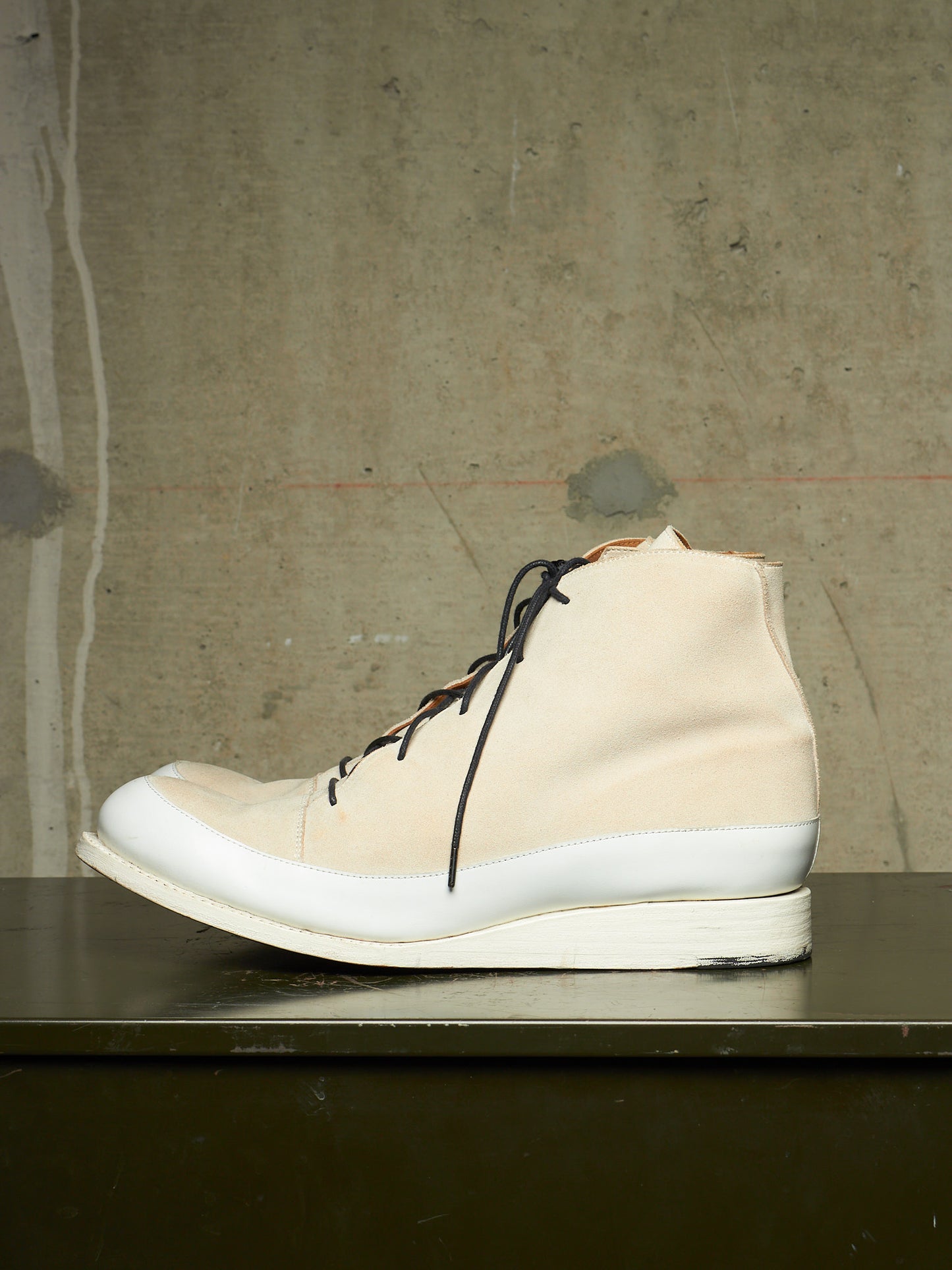 A high-top sneaker in off-white canvas with black laces and a thick white sole, resting on a dark surface against a textured concrete wall.