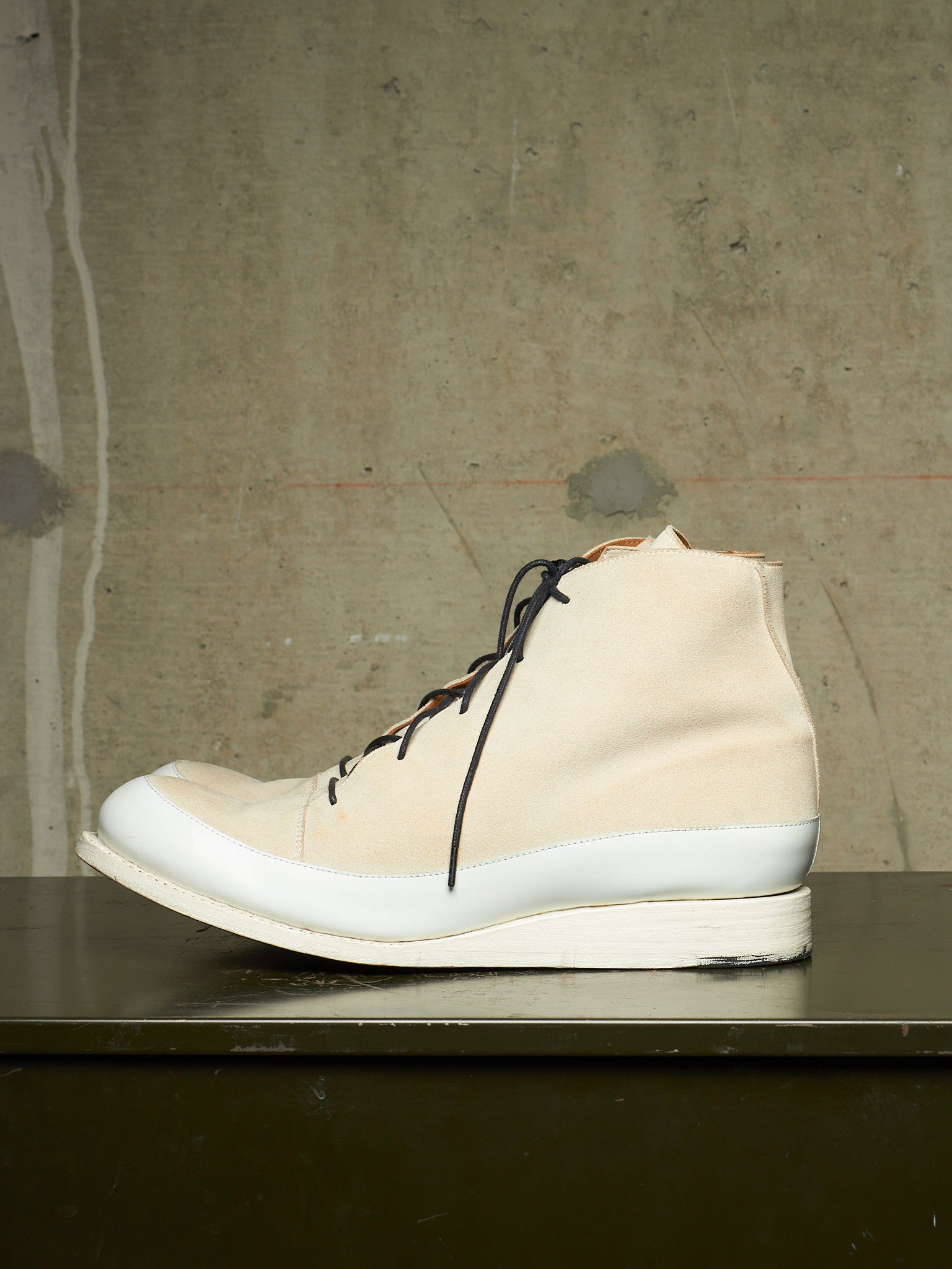 A high-top sneaker in off-white canvas with black laces and a thick white sole, resting on a dark surface against a textured concrete wall.
