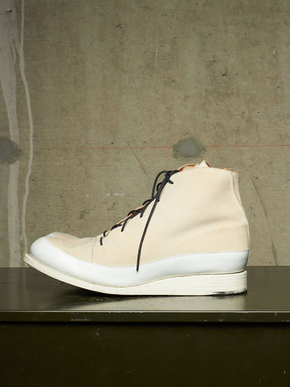 A high-top sneaker in off-white canvas with black laces and a thick white sole, resting on a dark surface against a textured concrete wall.