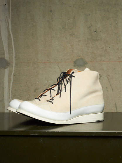 A single high-top sneaker in off-white canvas with black laces and a thick white sole, resting on a dark surface against a textured concrete wall.