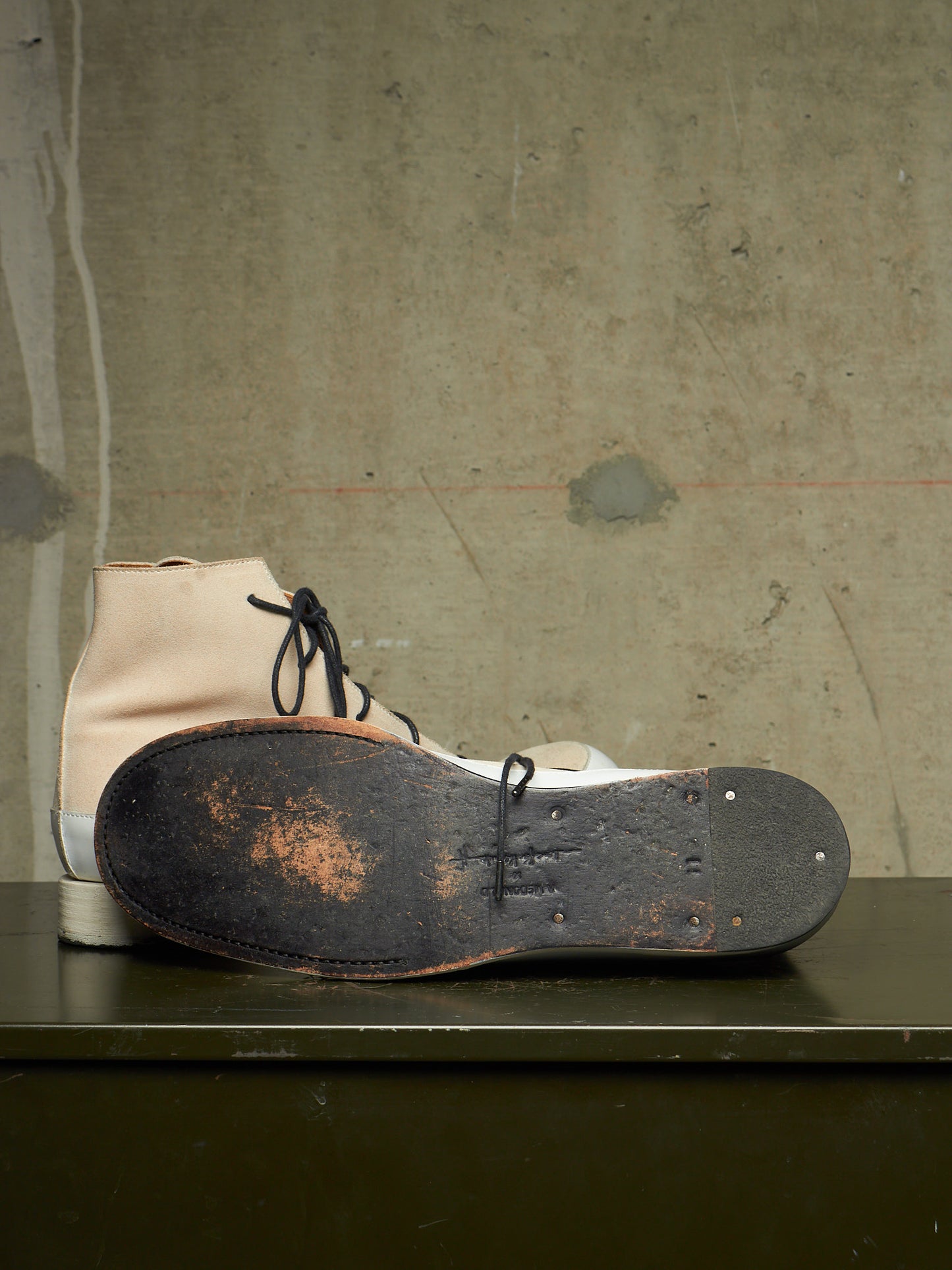 A pair of high-top boots with a cream-colored leather upper and white rubber sole, shown with one boot standing upright and the other lying on its worn black sole on a dark table against a textured concrete wall.