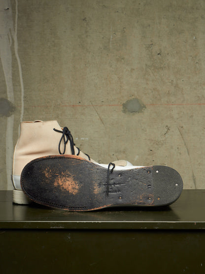 A pair of high-top boots with a cream-colored leather upper and white rubber sole, shown with one boot standing upright and the other lying on its worn black sole on a dark table against a textured concrete wall.