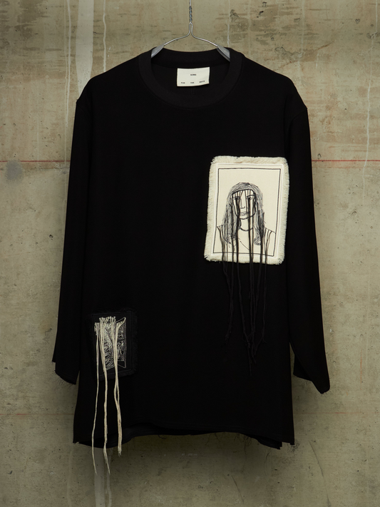 A black long-sleeve tunic top with distressed details, featuring a square white patch on the chest with a black line drawing of a face, and additional frayed white threads hanging from the lower left side, displayed on a hanger against a concrete wall.