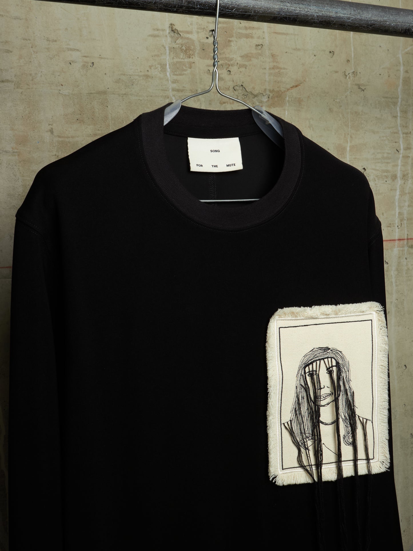 A black long-sleeve crewneck sweatshirt hanging on a metal hanger against a textured concrete wall. The sweatshirt features a square, raw-edge patch on the left chest containing a black line art illustration of a person with a headband.