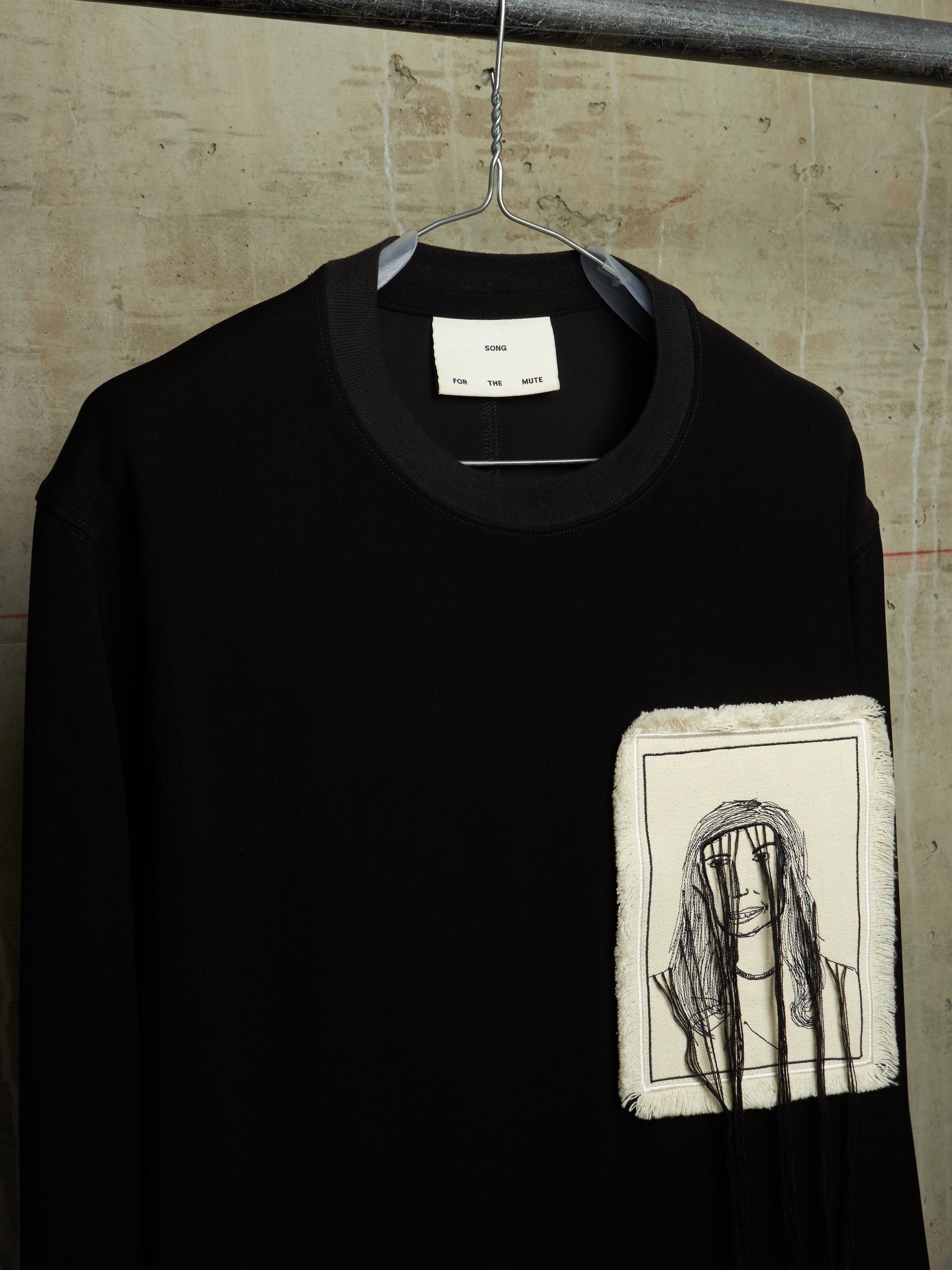 A black long-sleeve crewneck sweatshirt hanging on a metal hanger against a textured concrete wall. The sweatshirt features a square, raw-edge patch on the left chest containing a black line art illustration of a person with a headband.