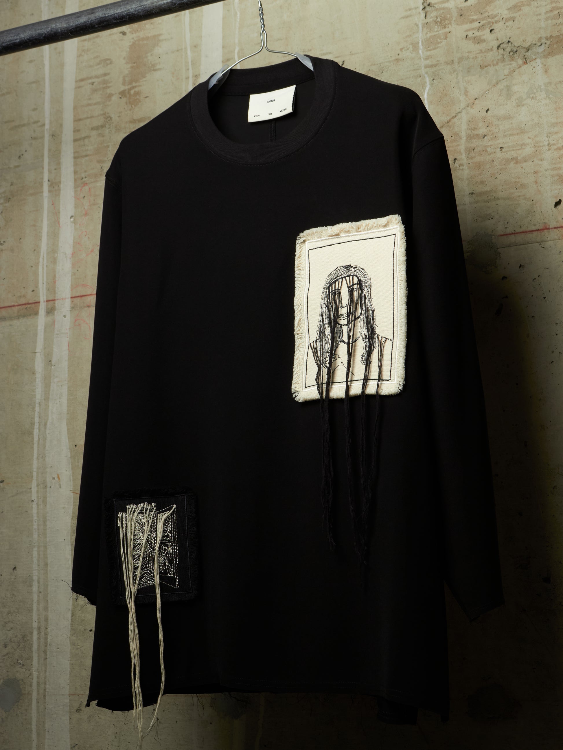 A black long-sleeve tunic top hanging on a hanger against a textured concrete wall. The shirt features a rectangular off-white patch on the chest with a black sketch of a face, and a smaller off-white patch on the lower left side with black text and hanging threads.
