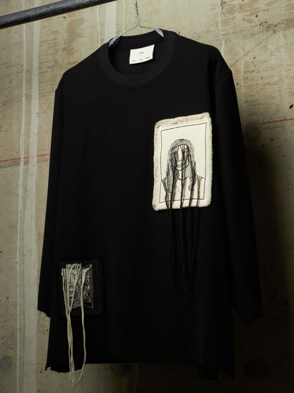 A black long-sleeve tunic top hanging on a hanger against a textured concrete wall. The shirt features a rectangular off-white patch on the chest with a black sketch of a face, and a smaller off-white patch on the lower left side with black text and hanging threads.