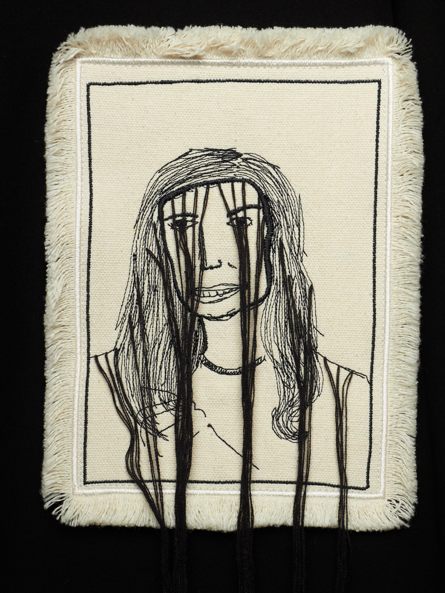 A square textile artwork or pillow cover with a cream woven background, black stitched portrait lines, and long black hanging threads in front, shown against a black background.