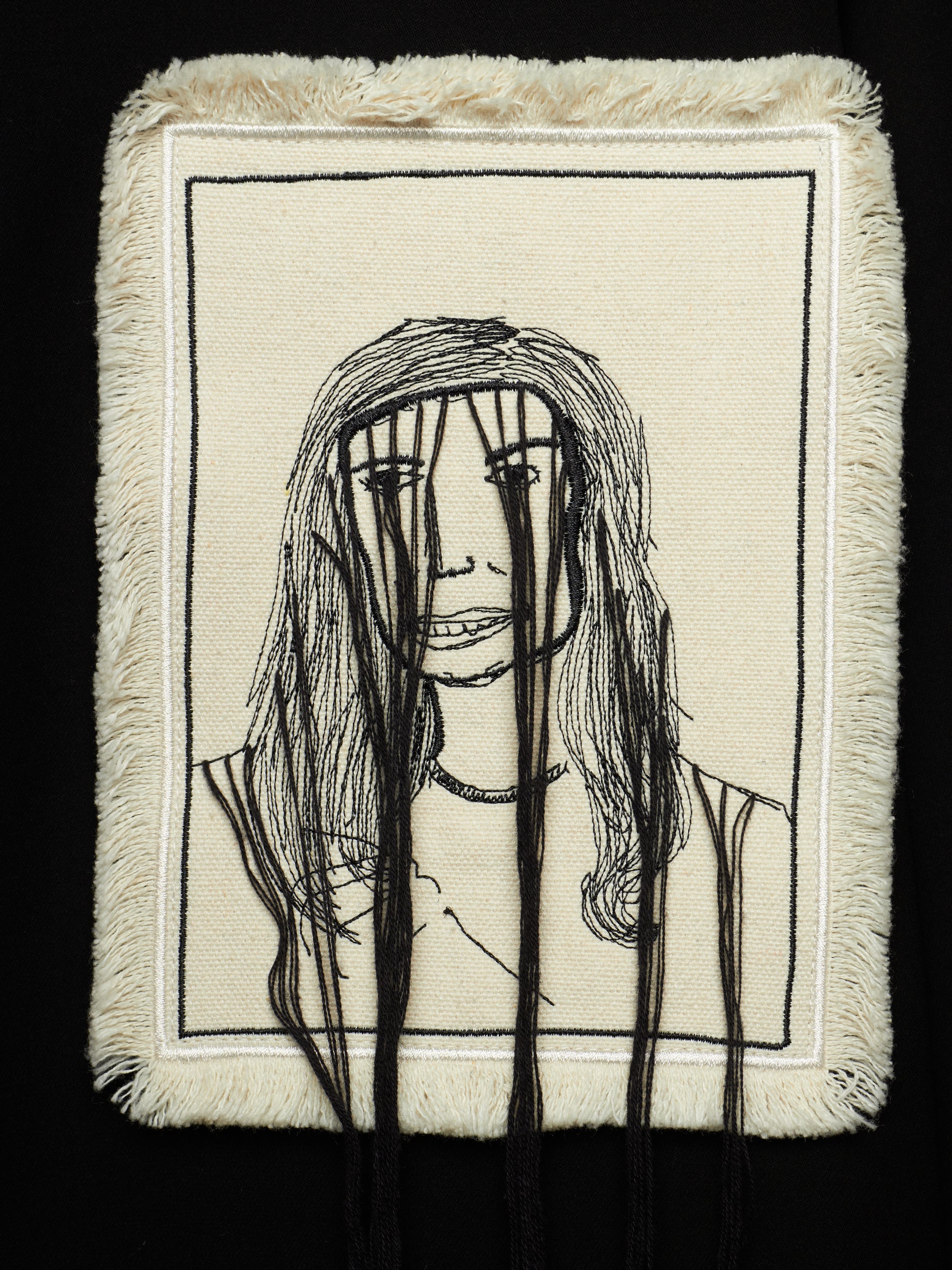 A square textile artwork or pillow cover with a cream woven background, black stitched portrait lines, and long black hanging threads in front, shown against a black background.