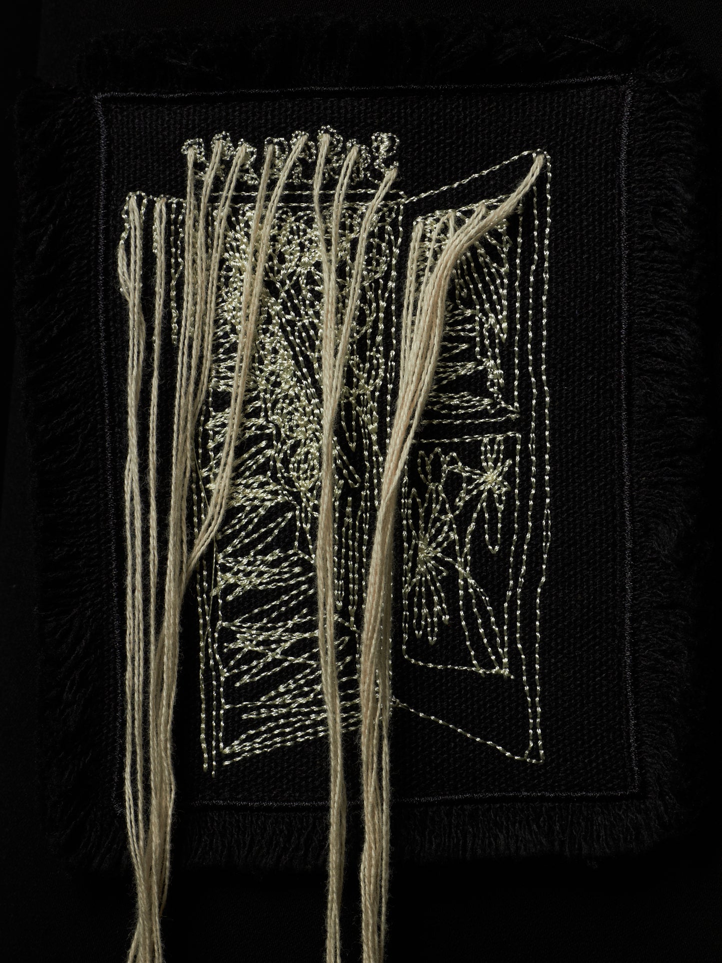 A square black textile artwork or patch featuring cream-colored thread embroidery and hanging strands, photographed against a black background.