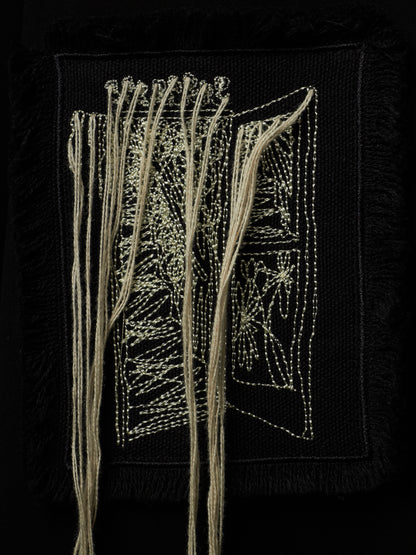 A square black textile artwork or patch featuring cream-colored thread embroidery and hanging strands, photographed against a black background.