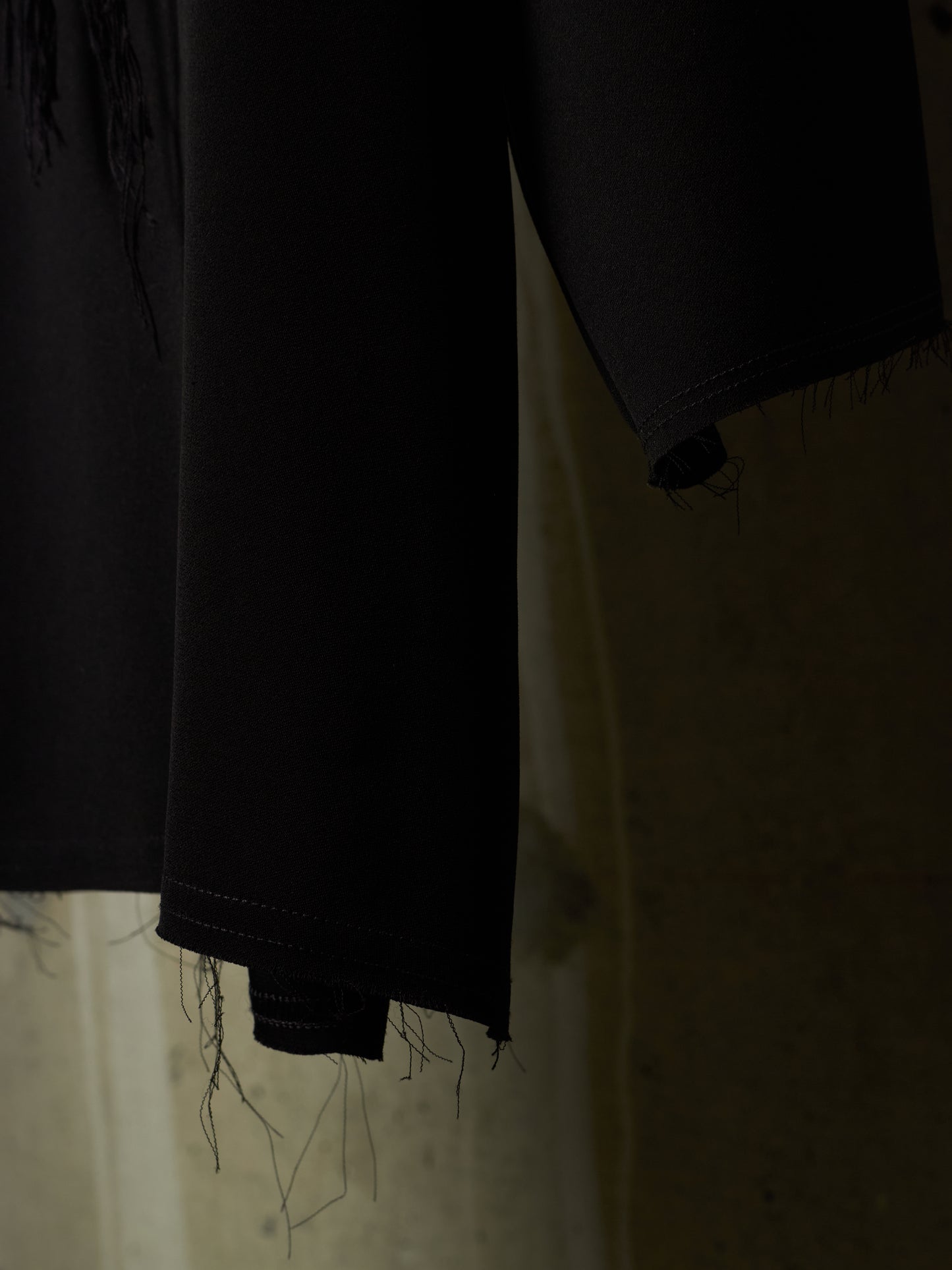 Close-up of a black garment with frayed, unfinished hems hanging against a dimly lit beige background.