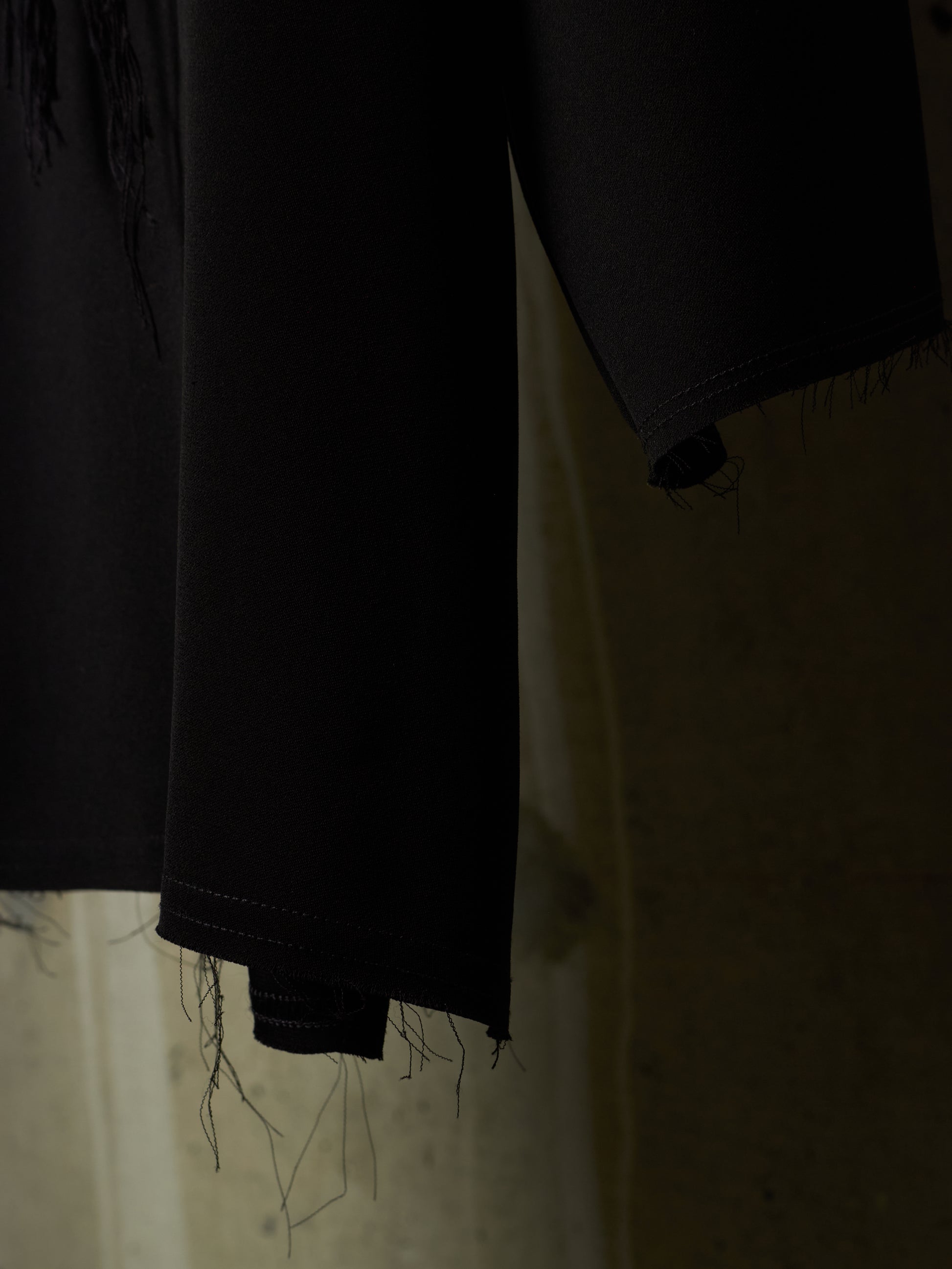 Close-up of a black garment with frayed, unfinished hems hanging against a dimly lit beige background.