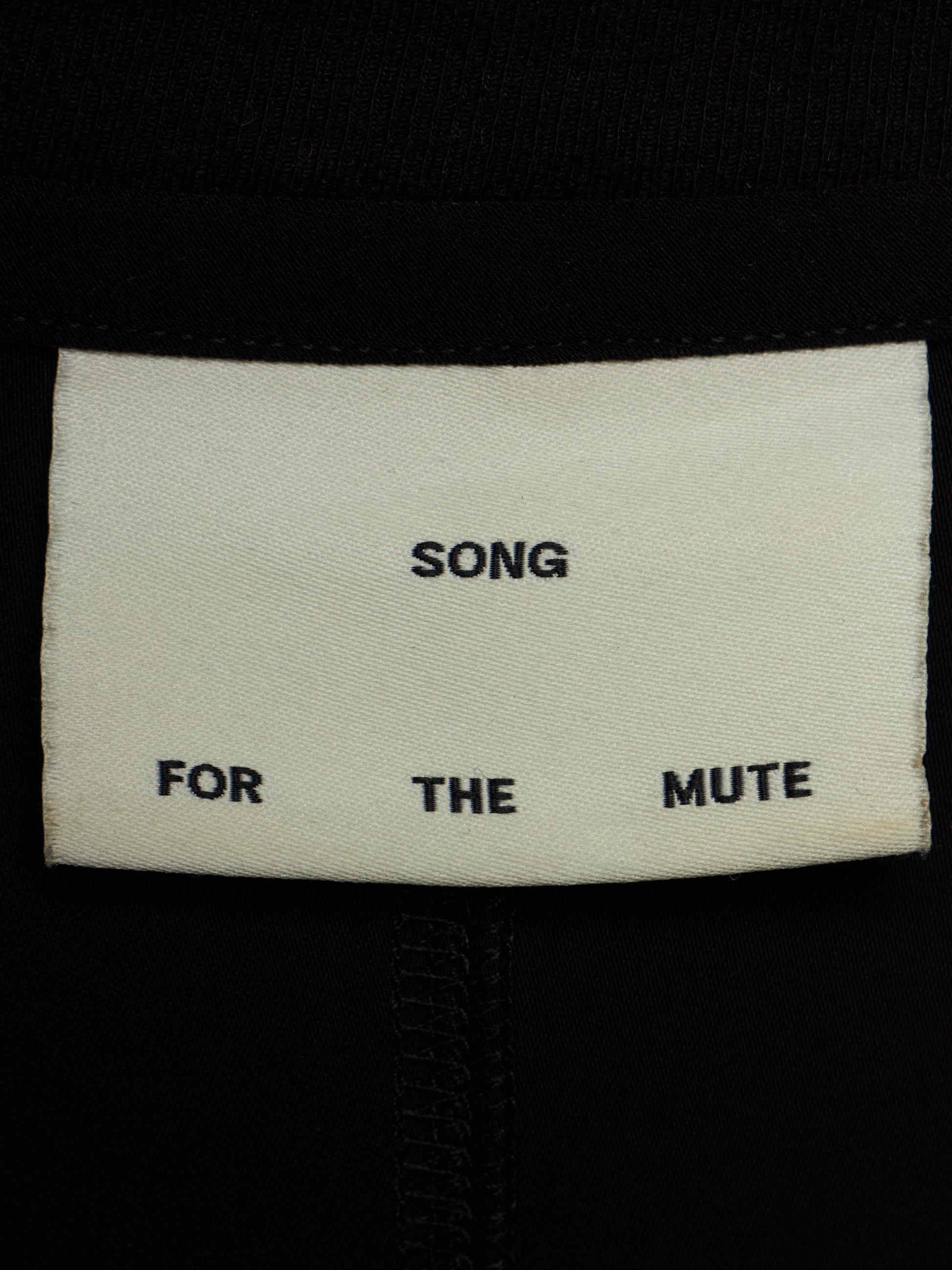 Close-up of a cream-colored fabric label sewn onto black clothing, printed with the text 'SONG FOR THE MUTE'.