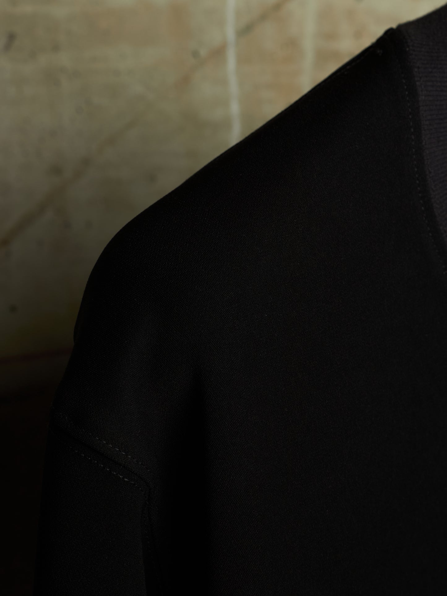 Close-up detail shot of a black knit garment, showing the shoulder and sleeve construction against a warm beige textured background.