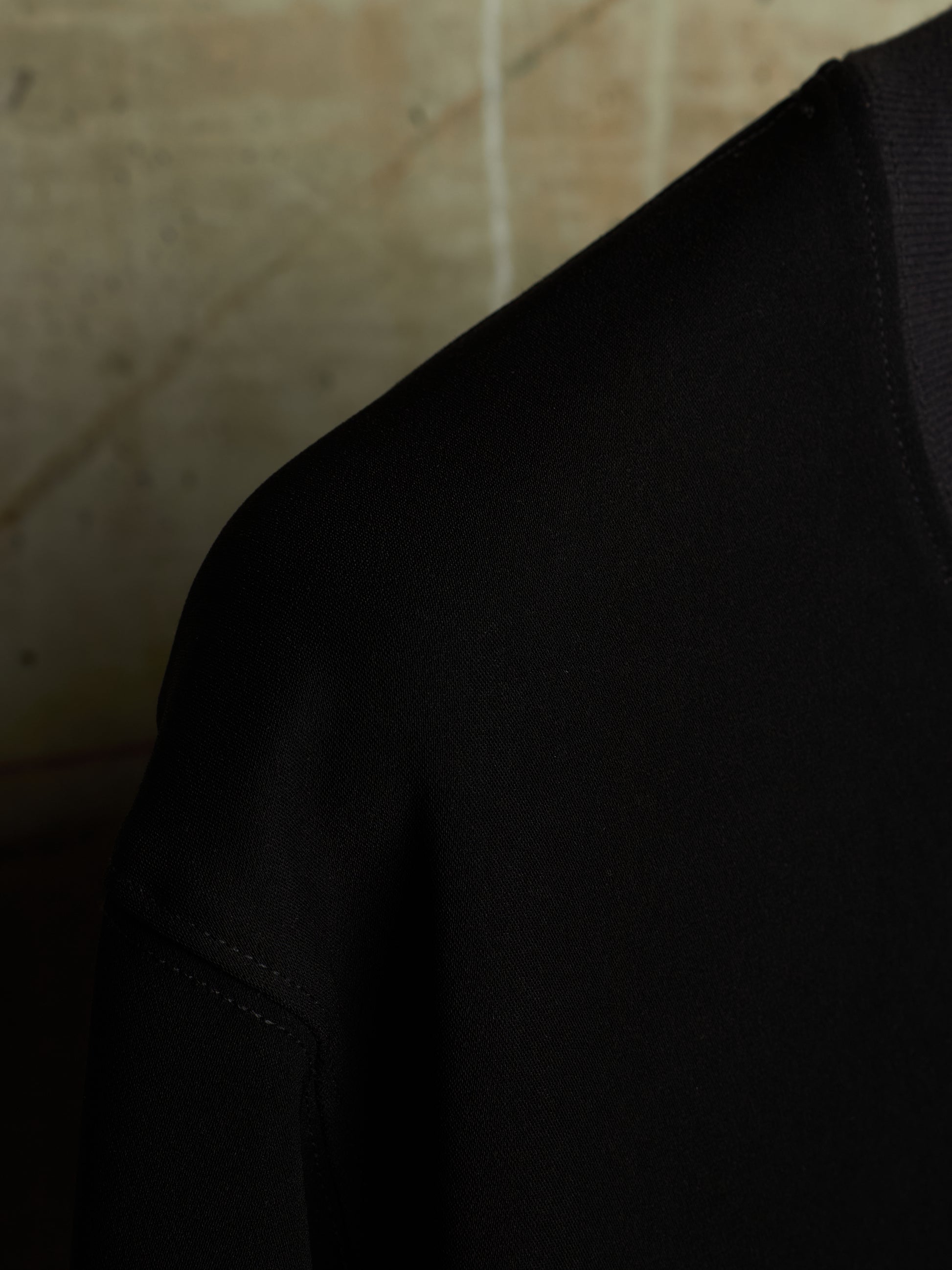 Close-up detail shot of a black knit garment, showing the shoulder and sleeve construction against a warm beige textured background.