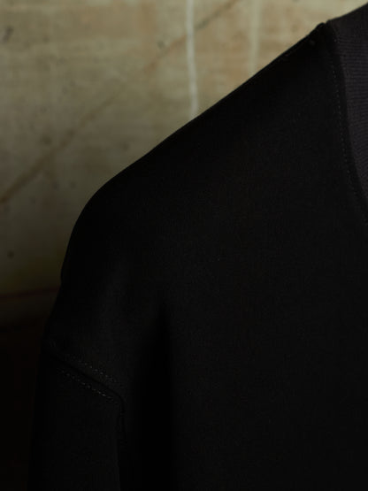 Close-up detail shot of a black knit garment, showing the shoulder and sleeve construction against a warm beige textured background.