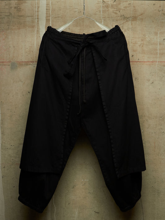Black wide-leg harem-style pants with a drawstring waist, hanging on a clear plastic hanger against a textured concrete wall.