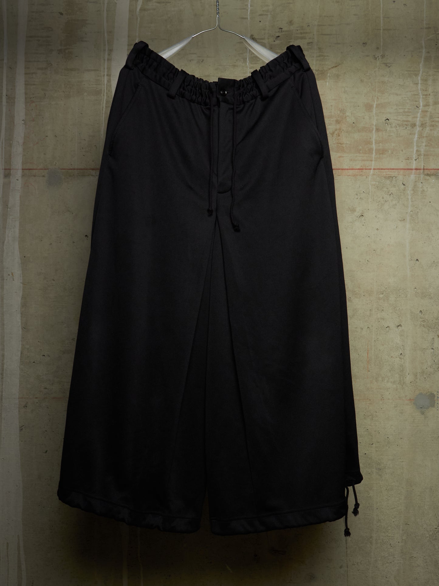 Black wide-leg cropped pants with an elastic drawstring waist, hanging on a white hanger against a textured concrete wall.