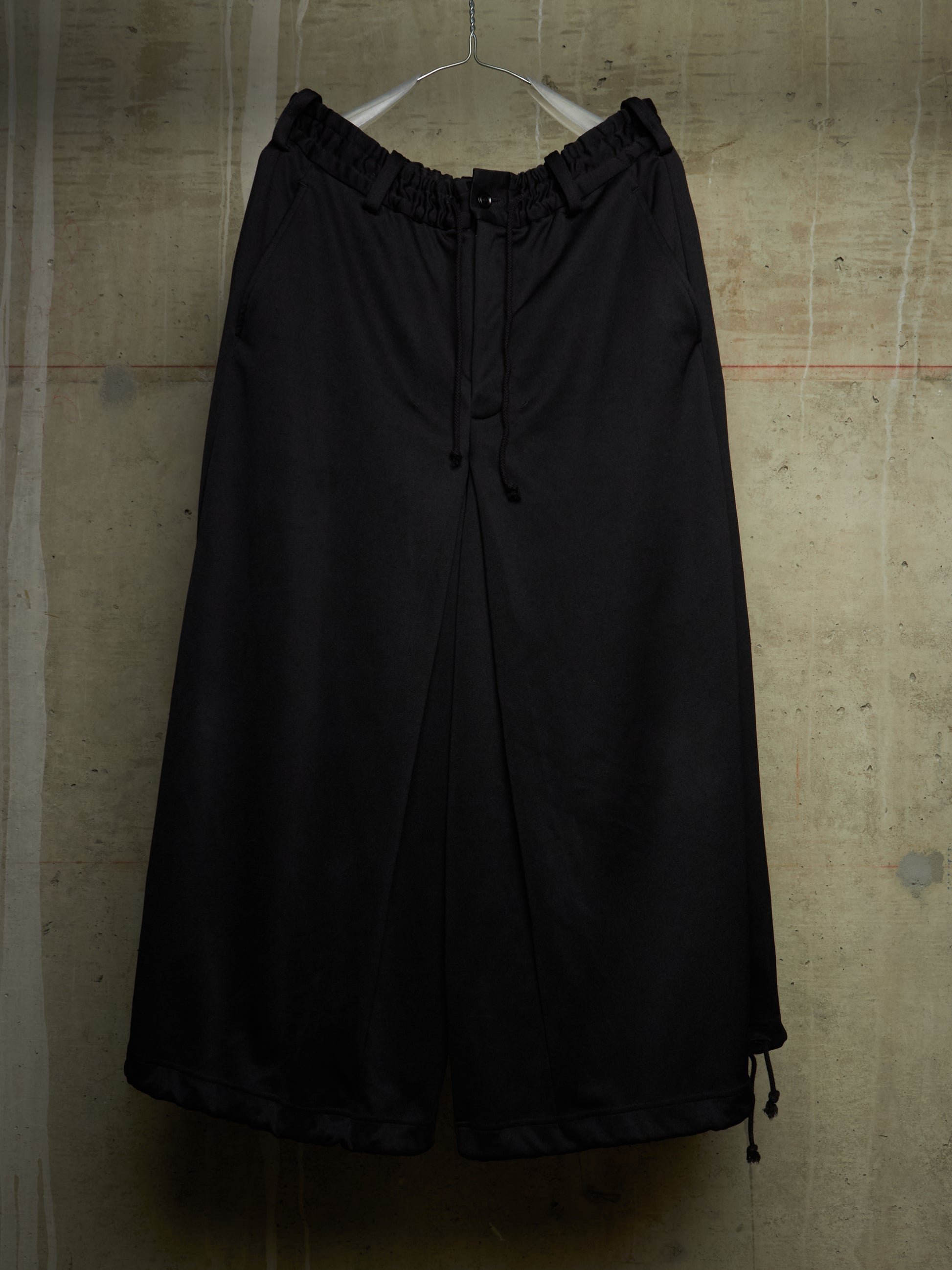 Black wide-leg cropped pants with an elastic drawstring waist, hanging on a white hanger against a textured concrete wall.