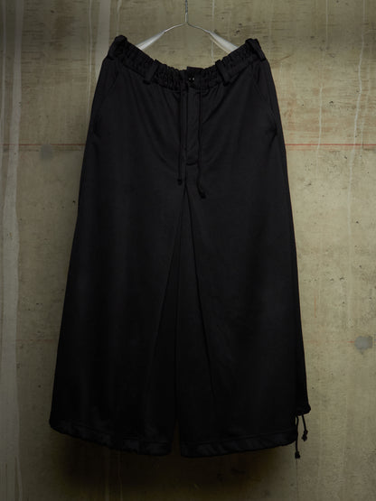 Black wide-leg cropped pants with an elastic drawstring waist, hanging on a white hanger against a textured concrete wall.