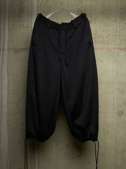 Black drawstring jogger pants with an elastic waistband and gathered ankle cuffs, hanging on a metal hanger against a textured concrete wall.