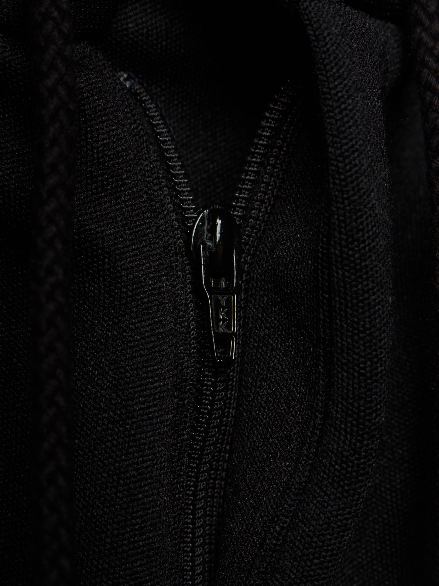 Close-up of a black zippered garment, showing the textured fabric and a black metal zipper pull with the brand name 'SAS' visible.