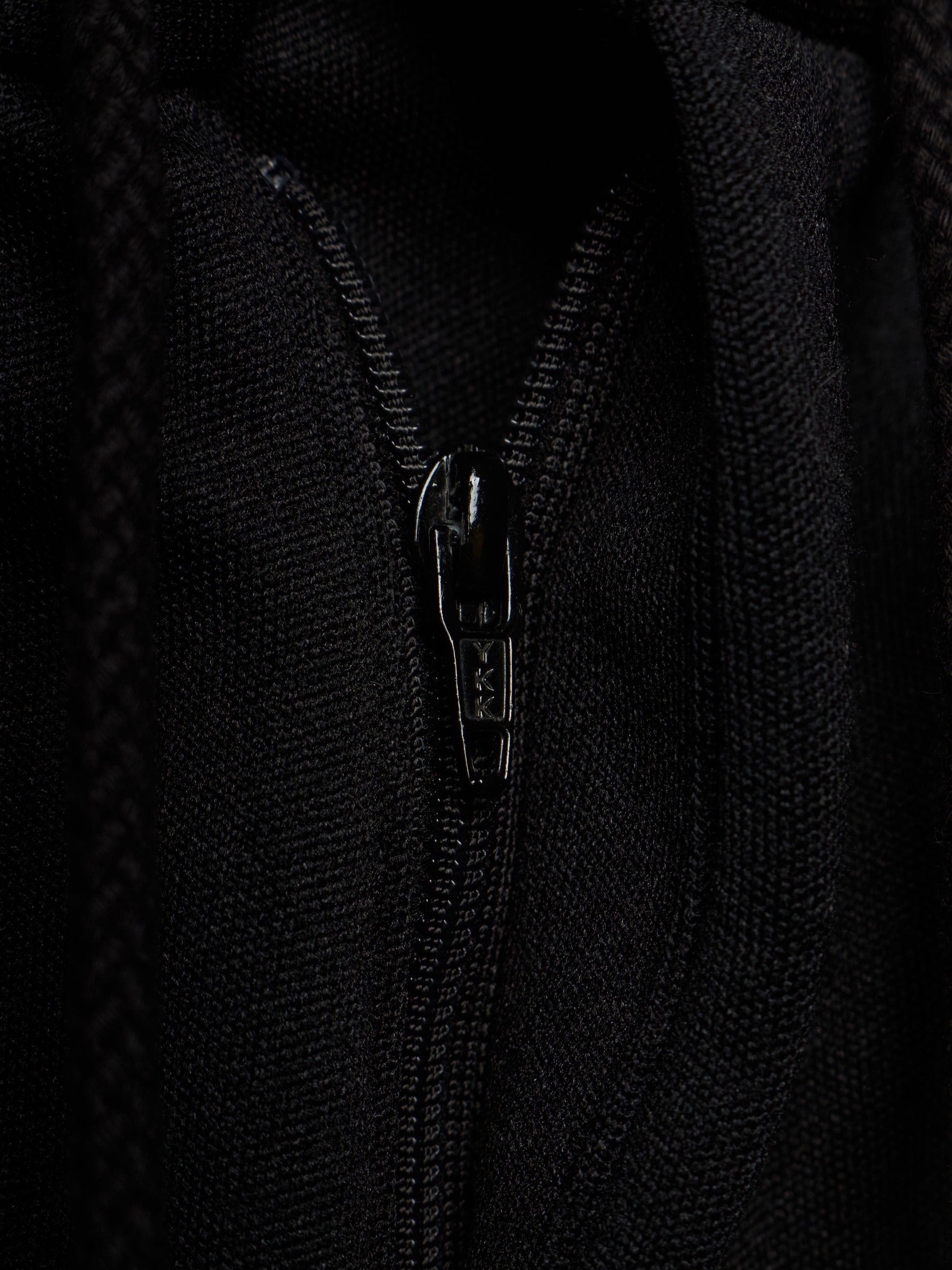 Close-up of a black zippered garment, showing the textured fabric and a black metal zipper pull with the brand name 'SAS' visible.