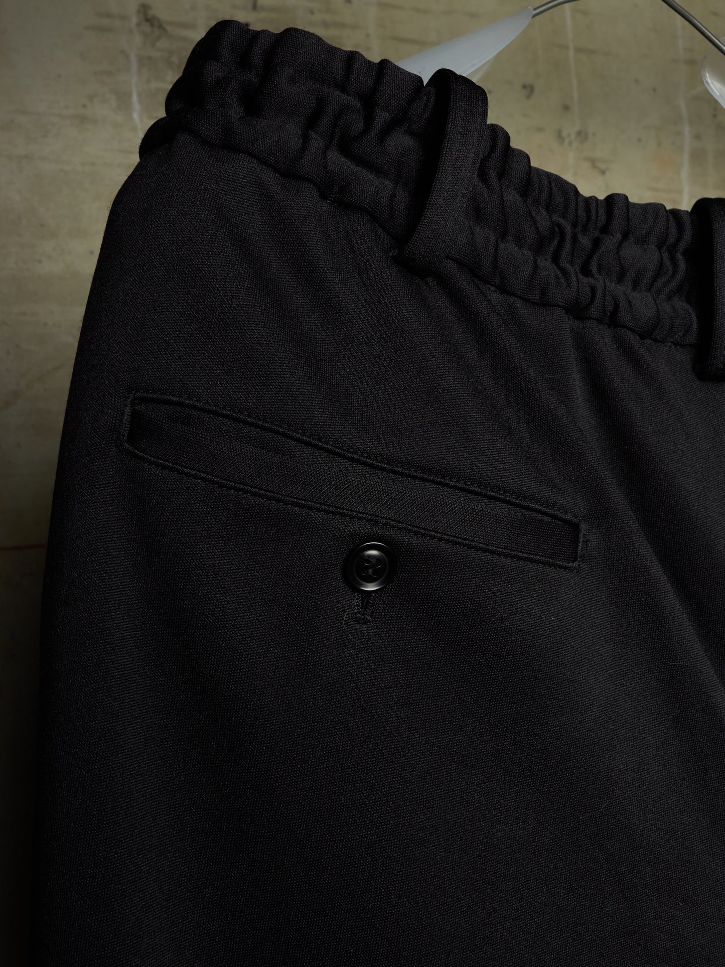 Close-up of black trousers featuring an elasticized waistband and a buttoned welt pocket, hanging against a neutral textured background.