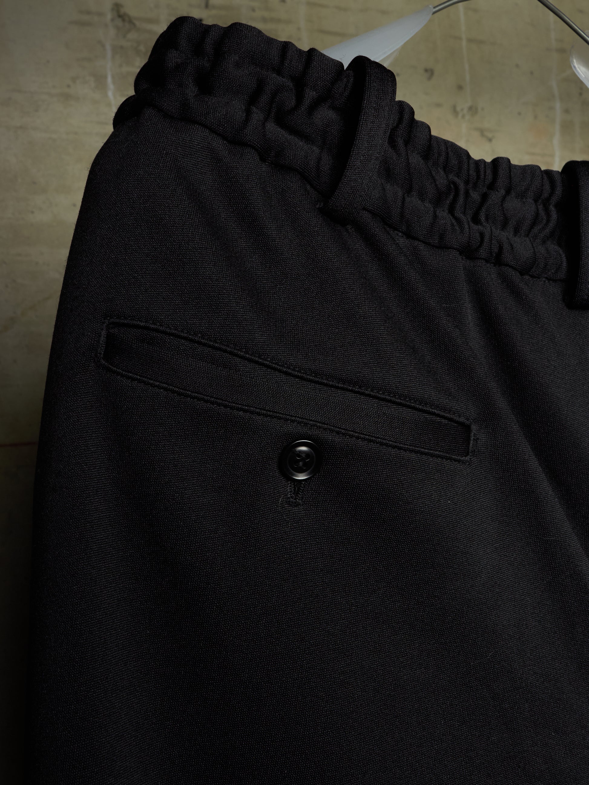Close-up of black trousers featuring an elasticized waistband and a buttoned welt pocket, hanging against a neutral textured background.